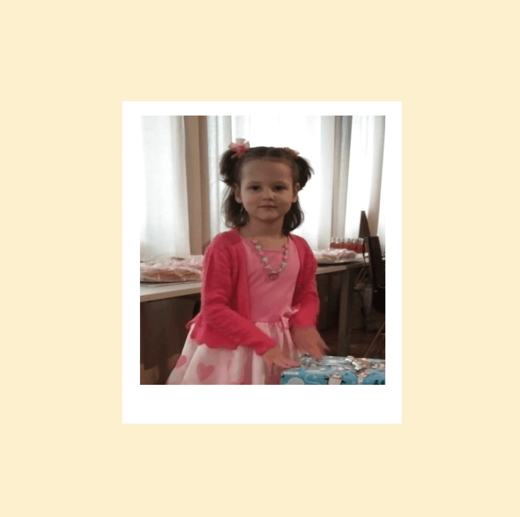 Young girl in a pink cardigan and dress standing indoors with gift boxes on a table.